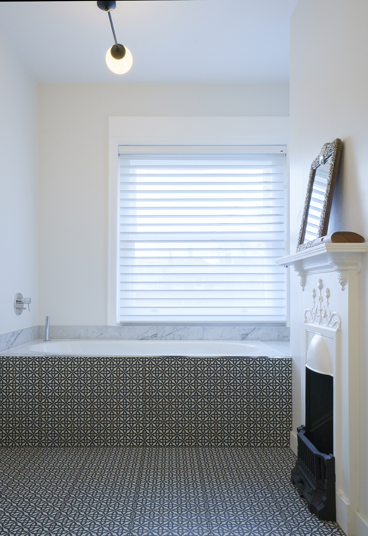 Contemporary bathroom renovation by Beacham Architects, featuring a built-in bathtub with patterned encaustic tiles, marble surround, restored Victorian fireplace, and soft natural light in a London home.